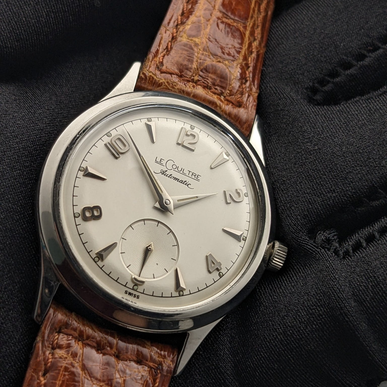 Jaeger-LeCoultre Automatic E328 displayed on a black glove, highlighting its light dial, Arabic numerals, and brown strap.